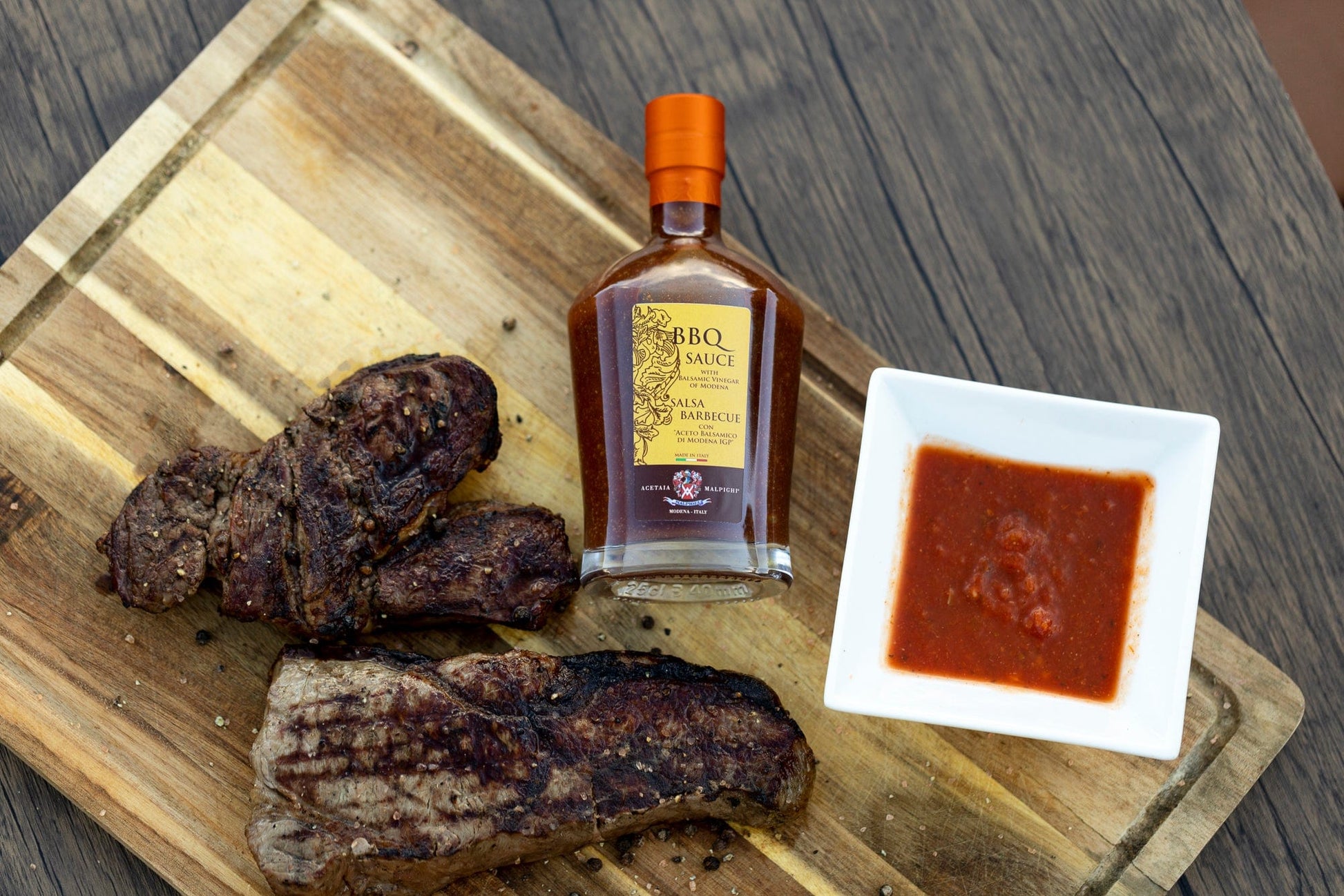 Bottle of BBQ sauce with a small dish of sauce and grilled meat on a wooden cutting board.