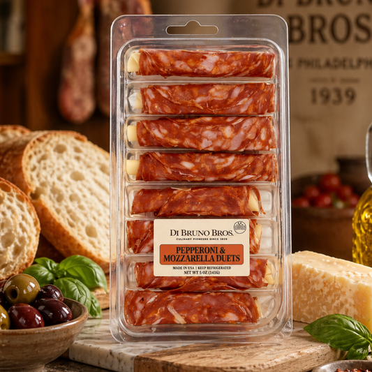 Packaged pepperoni and mozzarella bites with bread, olives, and basil on a wooden board.